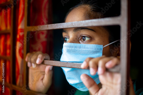 A bored Asian young girl wearing a protection surgical face mask at looking through window being in-home quarantine during coronavirus outbreak. Close-up views.