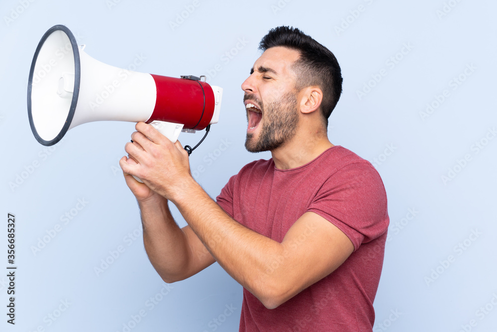 Young handsome man with beard over isolated blue background shouting through a megaphone Young handsome man with beard over isolated blue background shouting through a megaphone