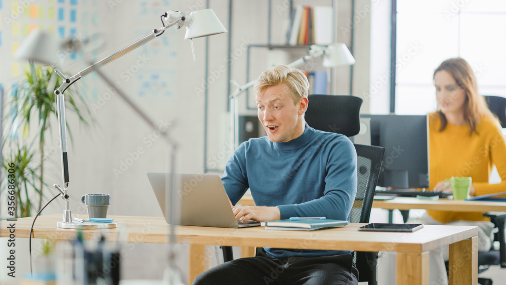 Handsome Caucasian IT Specialist Sitting at His Desk works on a Laptop with Surprised Face after Sudden Success in Project. Diverse Team of Young Professionals Working in Office. Handsome Caucasian IT Specialist Sitting at His Desk works on a Laptop with Surprised Face after Sudden Success in Project. Diverse Team of Young Professionals Working in Office.