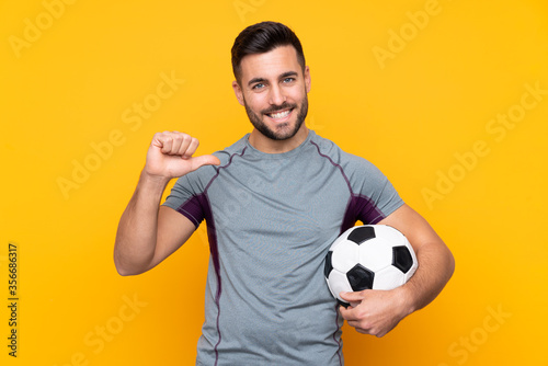 Man over isolated yellow background with soccer ball and proud of himself