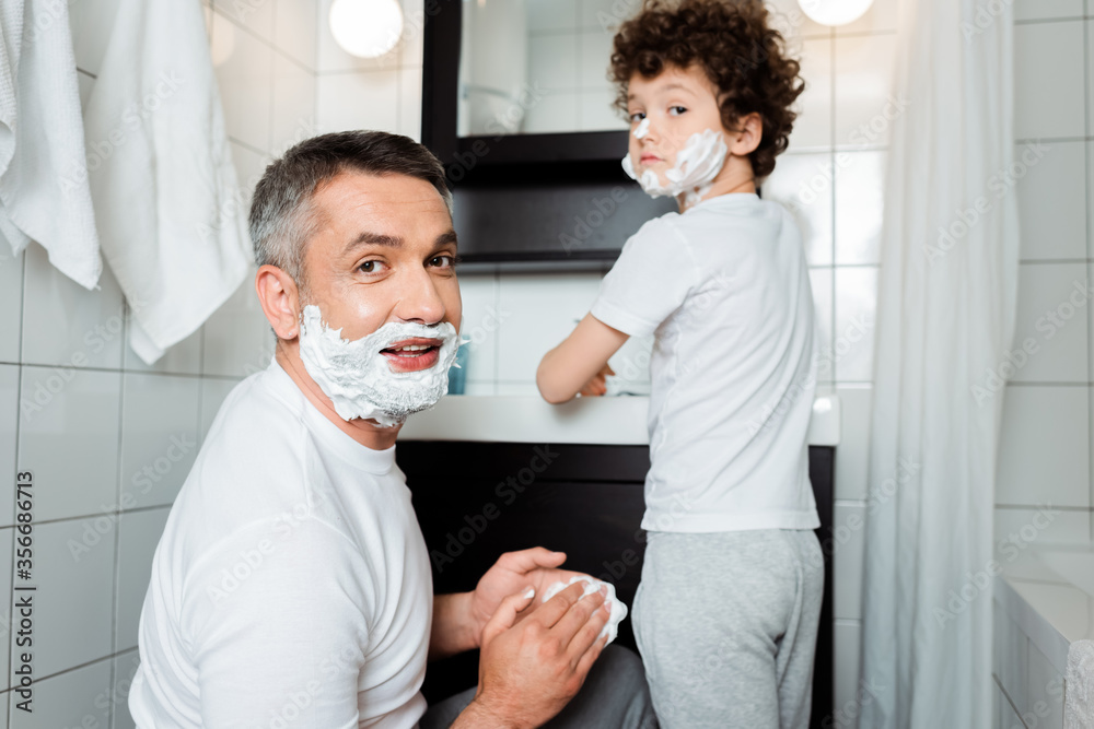 selective focus of father with shaving foam on face looking at camera near curly son in bathroom