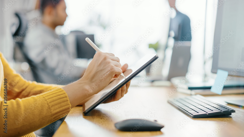 Anonymous Young Woman Sitting at Her Desk She's Drawing, Writing and Using Pen with Digital Tablet Computer. Focus on Hands with Pen. Bright Office where Diverse Team of Young Professionals Work. Anonymous Young Woman Sitting at Her Desk She's Drawing, Writing and Using Pen with Digital Tablet Computer. Focus on Hands with Pen. Bright Office where Diverse Team of Young Professionals Work.