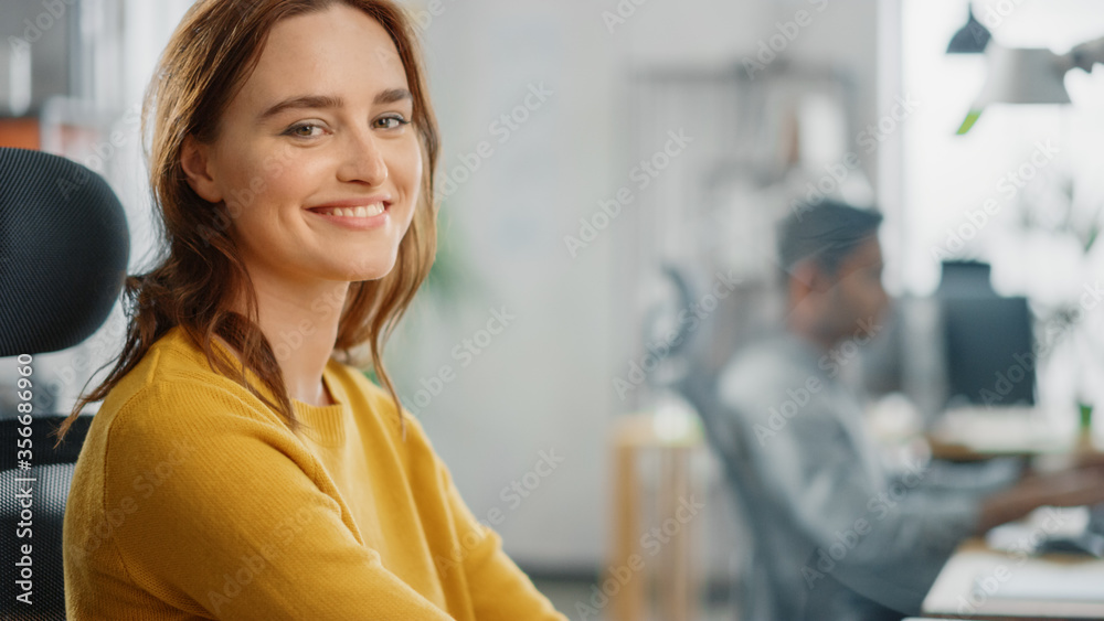 Portrait of Beautiful Young Woman with Red Hair Wearing Yellow Sweater Chats with Colleague and Turns Smilingly at Camera. Successful Woman Working in Bright Diverse Office.