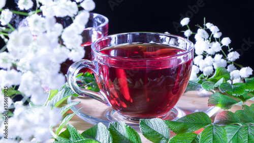 A mug of red tea in white hibiscus flowers and green leaves of medicinal tea on a wooden stand.Zen tea ceremony. Photo of red herbal Indian healing tea. Elegant mugs with a relaxing and tonic drink