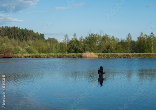 Fly Fisherman angler standing in calm water of forest lake, fish pond Kunraticky rybnik with birch and spruce trees growing along the shore and clear blue sky. Nature fishing background. Springtime
