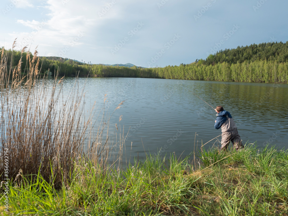 Fly Fisherman angler standing on shore of calm water of forest lake, fish pond Kunraticky rybnik with birch and spruce trees growing along the shore. Nature fishing background. Springtime