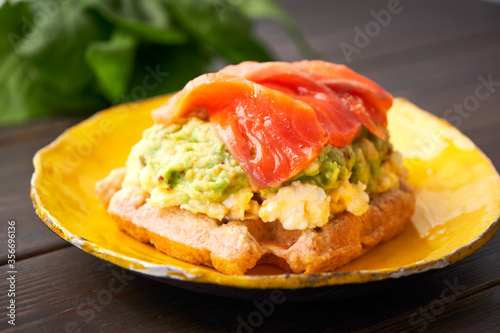 Belgian waffle with scrambled egg, avocado and salmon on wood table. Plate, bacon. Keto Breakfast variation.