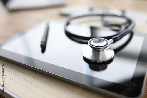 Close-up of modern black tablet device and doctors stethoscope. Silver pen on gadget. Macro shot medical equipment on desktop. Healthcare. Technology and medicine concept