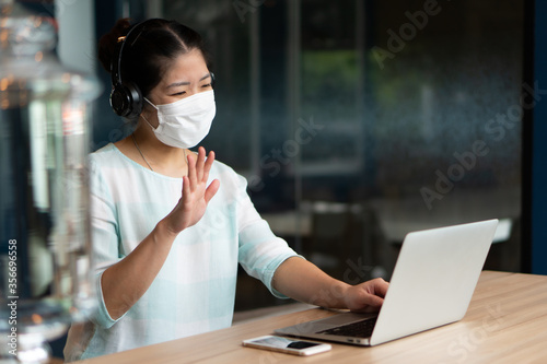 young asian woman wearing face mask and headphones and working remotely by using computer video call confernece at coworking space. social distancing and new normal lifestyle concept photo