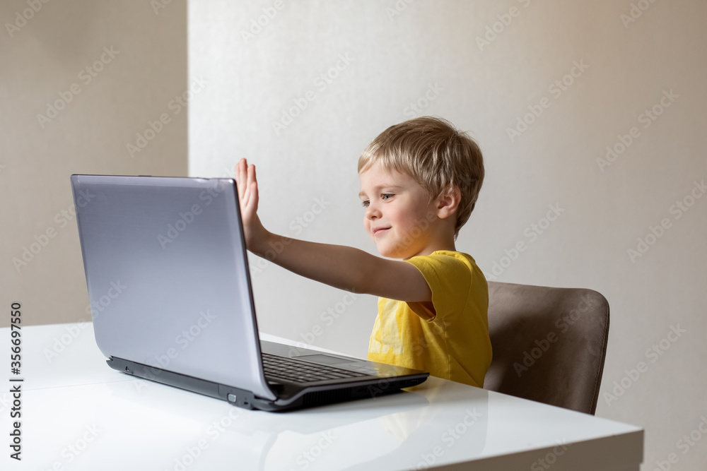 A cute little boy with blond hair sits at a table with a laptop and waves his hand. The boy smiles. He's got virtual communication. A cute little boy with blond hair sits at a table with a laptop and waves his hand. The boy smiles. He's got virtual communication.