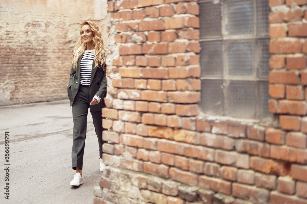 streetstyle photo of a beautiful woman with long blond curly hair who comes out of the corner and smiles, she is wearing a black suit, striped blouse and white sneakers, her hand in her pocket streetstyle photo of a beautiful woman with long blond curly hair who comes out of the corner and smiles, she is wearing a black suit, striped blouse and white sneakers, her hand in her pocket