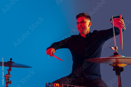 Caucasian male drummer improvising isolated on blue studio background in neon light. Performing, looks inspired, energy. Concept of human emotions, facial expression, ad, music, art, festival.