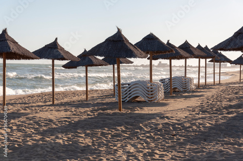 Thatched beach umbrellas, two piles of folded sunbeds on a deserted beach during quarantine against the background of the sea with waves.