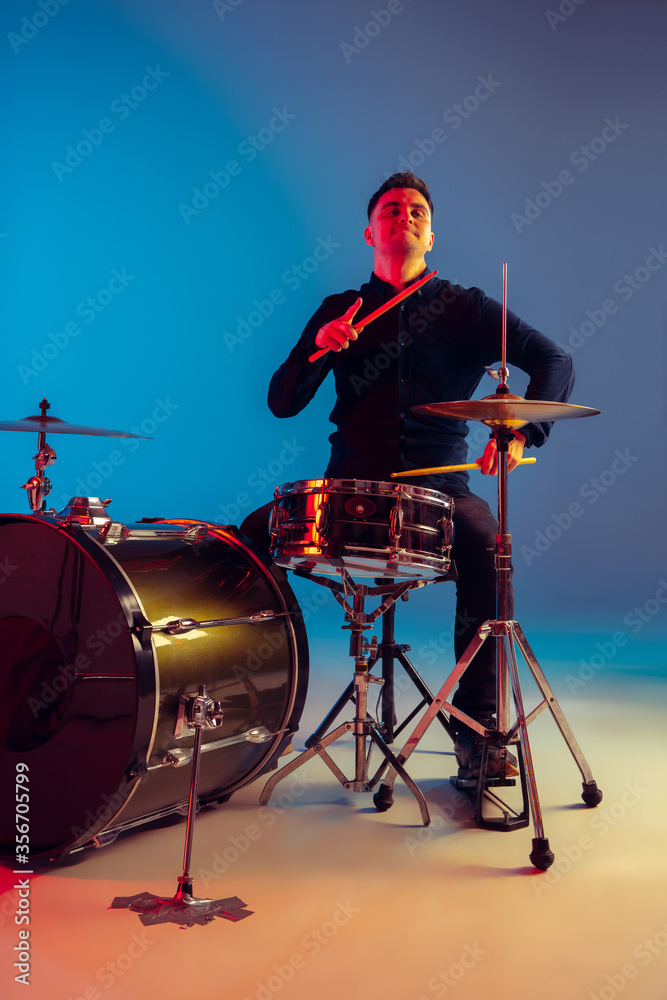 Fototapeta premium Caucasian male drummer improvising isolated on blue studio background in neon light. Performing, looks inspired, energy. Concept of human emotions, facial expression, ad, music, art, festival.