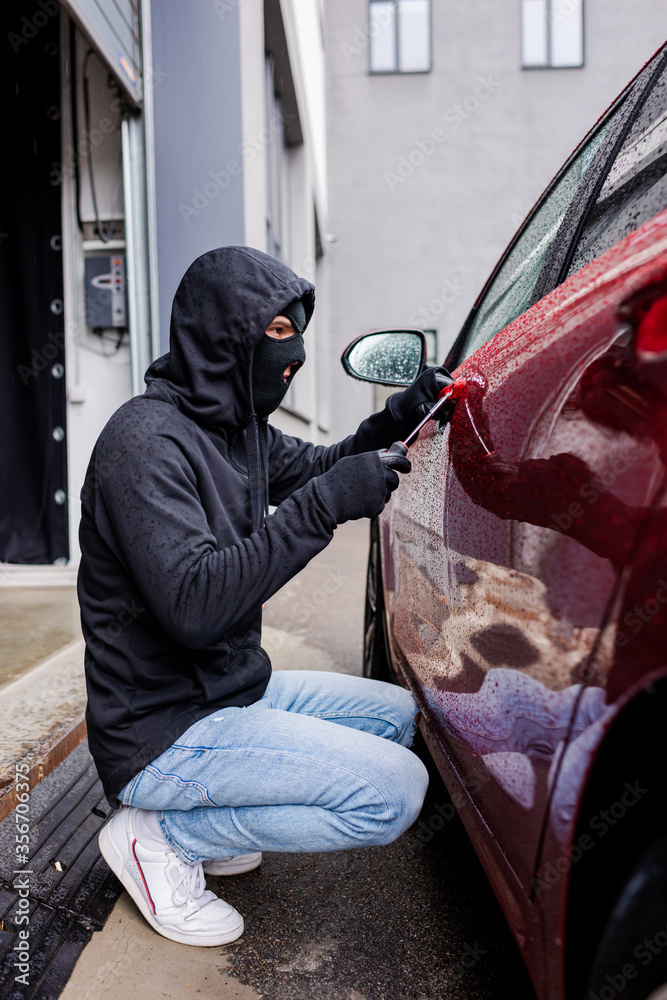 Side view of robber in balaclava using screwdriver while opening lock on door of car on urban street Side view of robber in balaclava using screwdriver while opening lock on door of car on urban street
