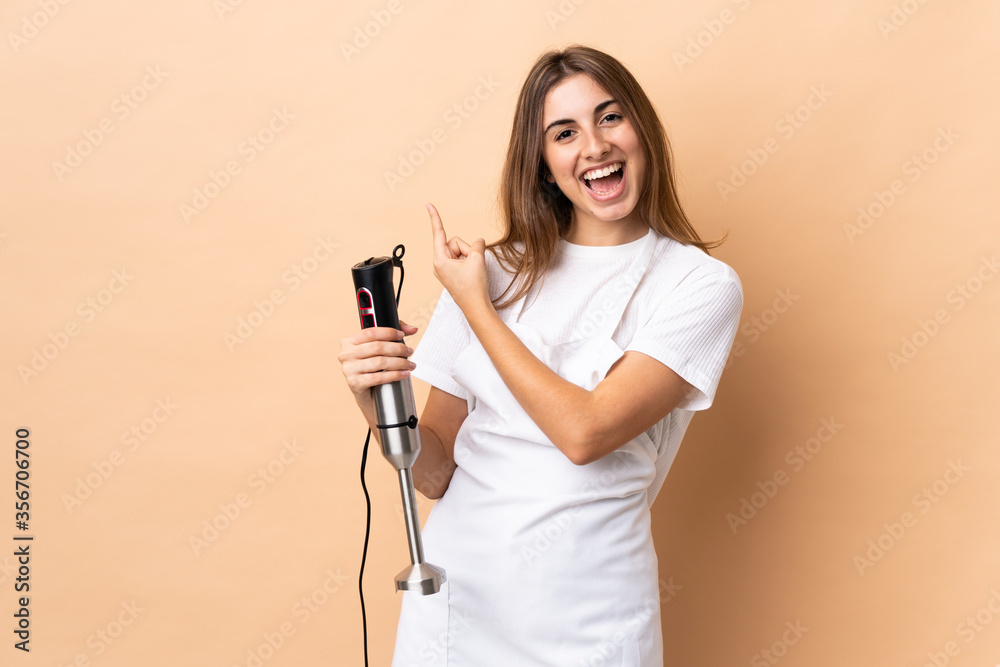 Woman using hand blender over isolated background surprised and pointing side Woman using hand blender over isolated background surprised and pointing side