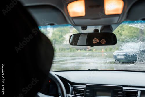 Selective focus of man in balaclava looking at mirror in car