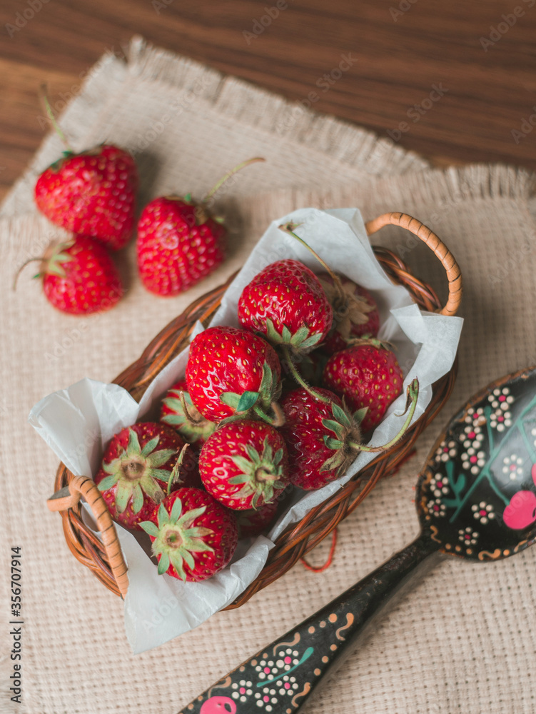 Close-up view of strawberries in wicker bowl on table. Mellow strawberries in basket on tablecloth with wooden painted spoon Close-up view of strawberries in wicker bowl on table. Mellow strawberries in basket on tablecloth with wooden painted spoon