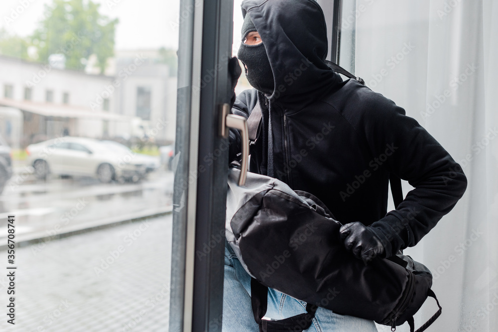 Robber in balaclava and leather gloves holding bag near open window