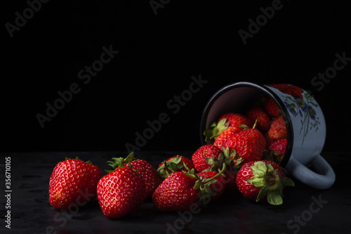 Strawberries on a black background. An enameled mug lies on a table with strawberries in it. Red strawberries. Creative photo of strawberries