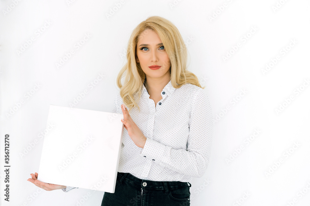 Beautiful blonde woman on a light background in a white shirt holds a white sheet on the left Beautiful blonde woman on a light background in a white shirt holds a white sheet on the left
