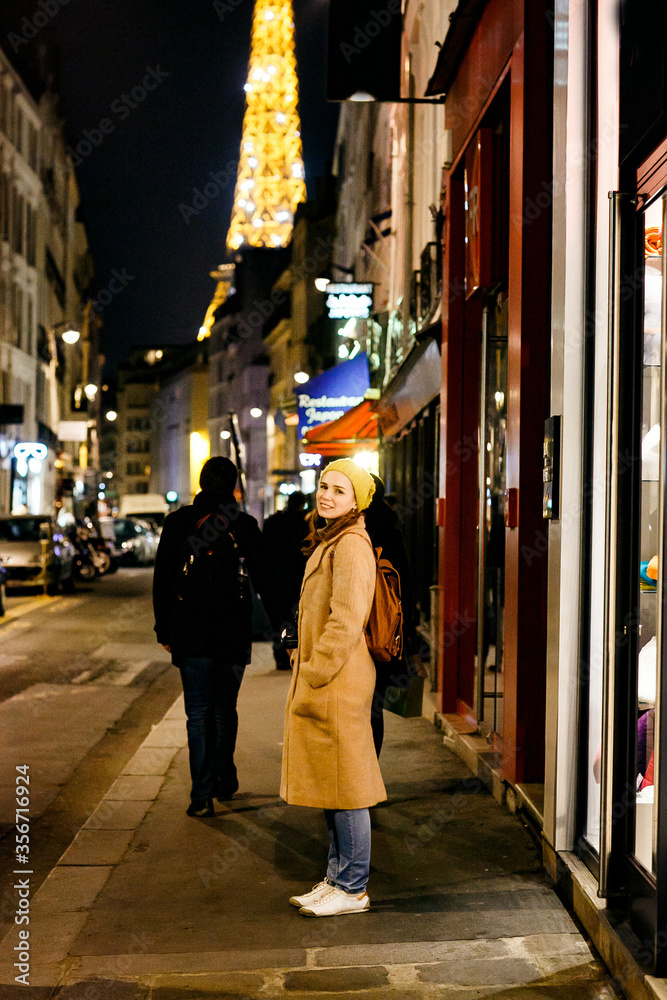 woman in trench coat with backpack walking in night Paris.