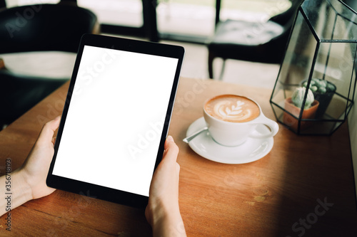 A woman hands are holding a mockup of black tablet with blank white screen in the cafe with a white cup of latte coffee. sitting on the wood table, clipping path included