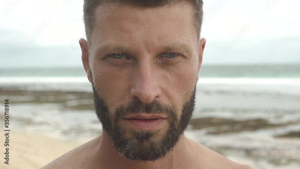 Close-up of a young handsome man with beard looks at the camera on background of the ocean Close-up of a young handsome man with beard looks at the camera on background of the ocean