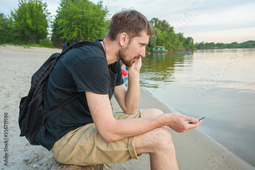 Young male tourist talking on a cell phone, on the river bank. Lifestyle, side view.