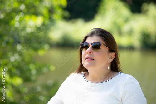 Portrait of a lady outdoors. She is caucasian in her 40s. She is wearing sunglasses and is enjoying the view of the park.