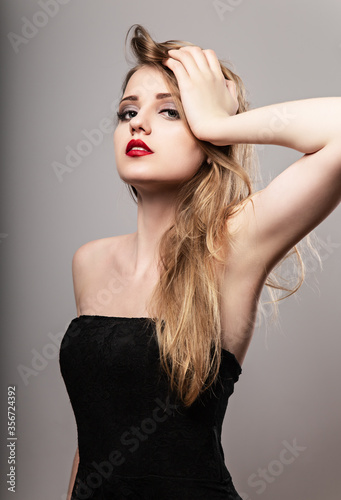 Beautiful bright makeup woman with long blond hair style in black dress, red lipstick looking with epilation armpit and sexy look on grey background.. Closeup