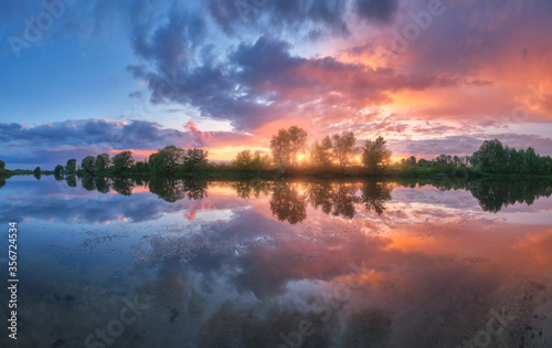 Beautiful river coast at sunset in summer. Colorful landscape with lake, green trees and grass, blue sky with multicolored clouds and orange sunlight reflected in water. Nature. Vibrant scenery © den-belitsky