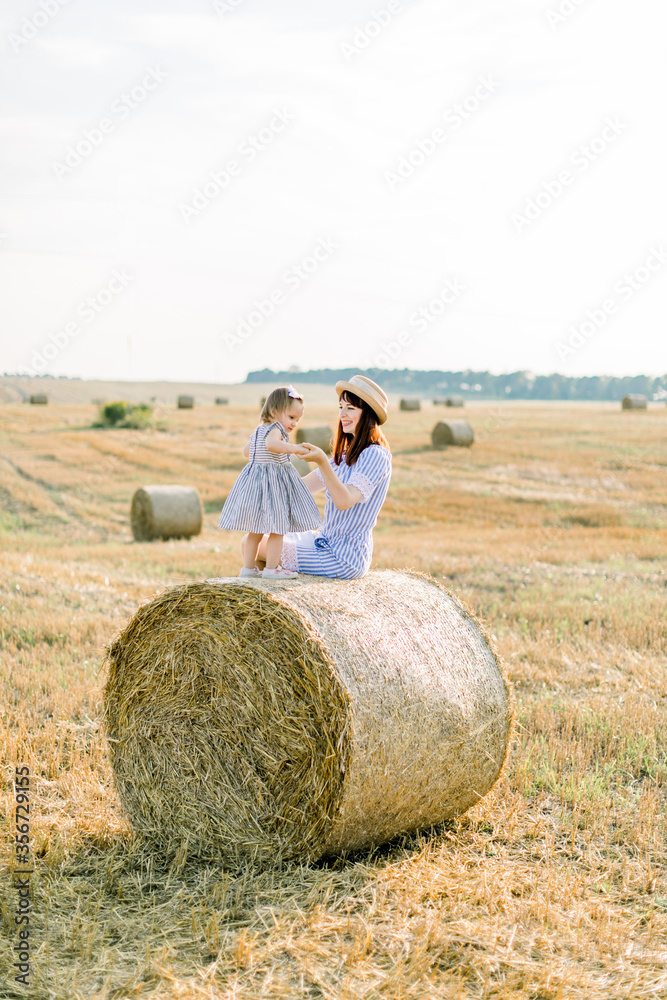 Happy two year old girl child in striped dress playing with young smiling mom in summer autumn field with hay bales, sitting together on hay stack. Summer sunset portrait Happy two year old girl child in striped dress playing with young smiling mom in summer autumn field with hay bales, sitting together on hay stack. Summer sunset portrait