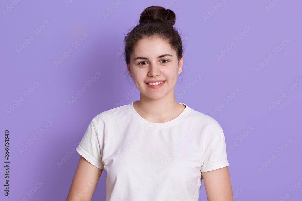 Sensual young woman with hair bun against lilac background. Attractive female model with perfect skin and charming smile, wearing white casual t shirt, teenage with positive emotions. Sensual young woman with hair bun against lilac background. Attractive female model with perfect skin and charming smile, wearing white casual t shirt, teenage with positive emotions.