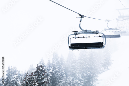 An empty chairlift appearing through the foggy cloud layer on a mountainside. Snow covered trees are seen below. 