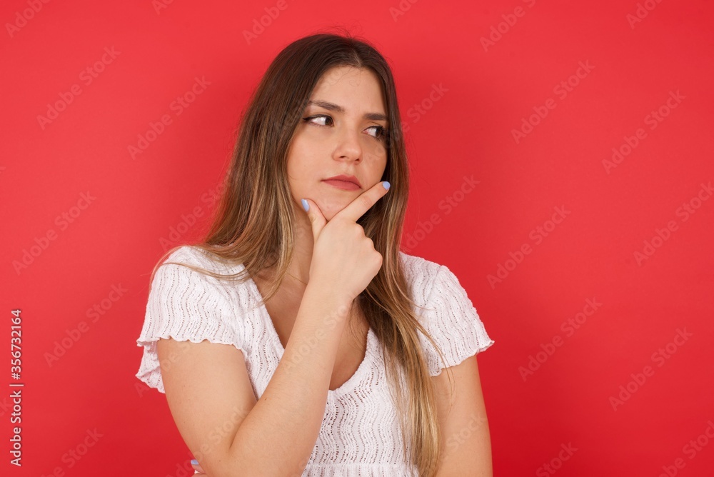 Isolated portrait of stylish young European woman with hand under chin and looking sideways with doubtful and skeptical expression, worry and doubt. Standing indoors over gray background. Isolated portrait of stylish young European woman with hand under chin and looking sideways with doubtful and skeptical expression, worry and doubt. Standing indoors over gray background.