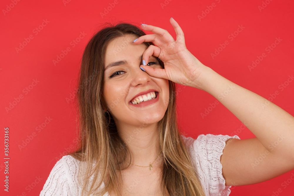 Beautiful young blonde woman with happy face smiling doing ok sign with hand on eye looking through finger. Wearing casual clothes and standing. Beautiful young blonde woman with happy face smiling doing ok sign with hand on eye looking through finger. Wearing casual clothes and standing.