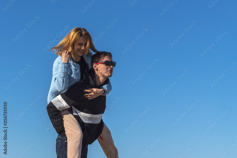 Happy man carry pickaback his girlfriend on blue sky background. Young couple enjoying travel outdoors. Love story of attractive, cheerful, funny couple. Happy man carry pickaback his girlfriend on blue sky background. Young couple enjoying travel outdoors. Love story of attractive, cheerful, funny couple.