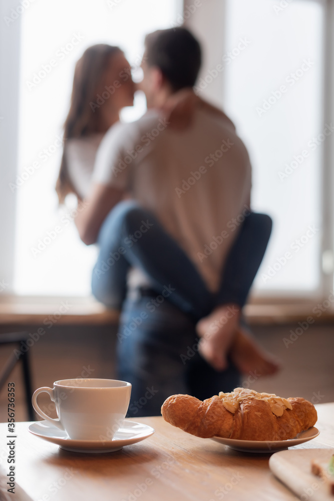 selective focus of cup of coffee near croissant and couple in kitchen