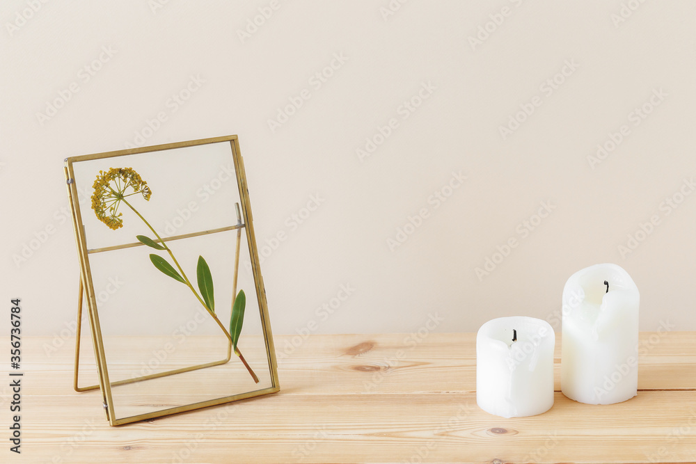 Composition of golden metal photo frame with yellow dried flower and candles on a wooden table on pastel beige background. Stylish home interior decor. Modern interior design concept. Composition of golden metal photo frame with yellow dried flower and candles on a wooden table on pastel beige background. Stylish home interior decor. Modern interior design concept.