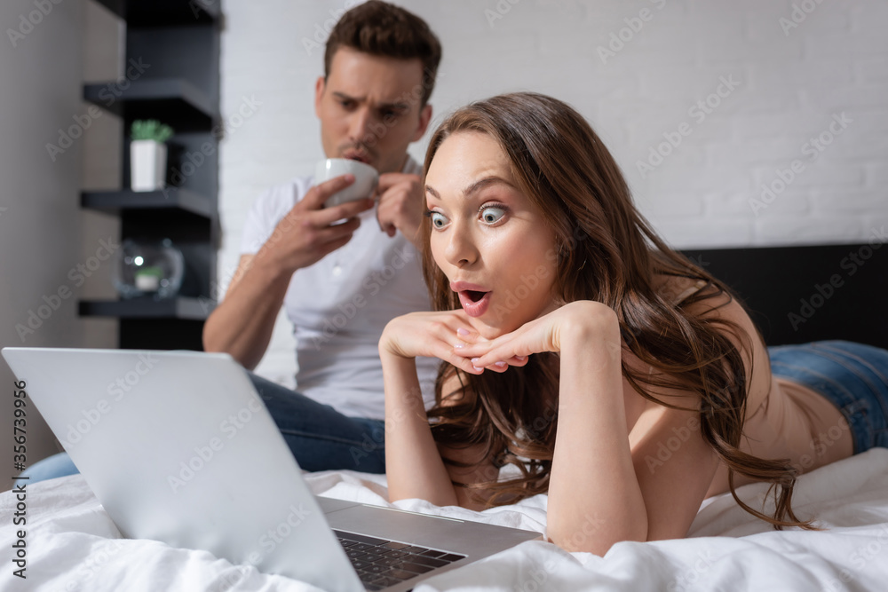 selective focus of surprised woman watching movie on laptop near boyfriend with cup selective focus of surprised woman watching movie on laptop near boyfriend with cup