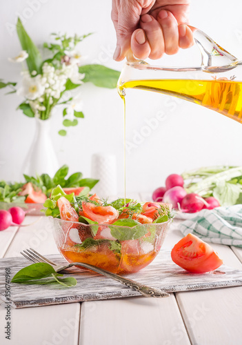 Woman pours oil into a salad with vegetables and herbs in a transparent salad bowl on a white wooden background.