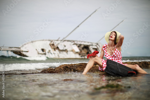 Woman in red dress with red lipstick escaped from sunken yacht.She is holding red women shoes and near her black suitcase.