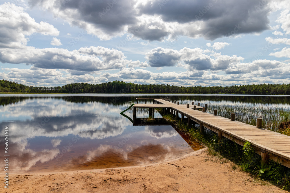 Landscape with a swamp lake and with a wooden footbridge. Beautiful clouds and the surrounding forest were reflected in dark water. Latvia. Lake Ratnieku