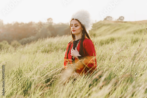 Georgian girl with closed eyes in white papakha and red national dress seats on the green grass and dreams. Georgian culture lifestyle. photo