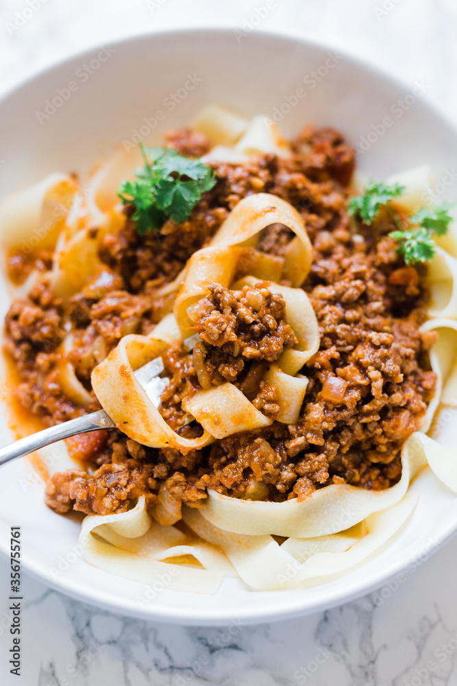 A fork is full of freshly homemade pappardelle pasta noodles in a meat bolognese sauce. This is Italian home cooking. The spaghetti bolognese is served in a white bowl and garnished with parsley. A fork is full of freshly homemade pappardelle pasta noodles in a meat bolognese sauce. This is Italian home cooking. The spaghetti bolognese is served in a white bowl and garnished with parsley.