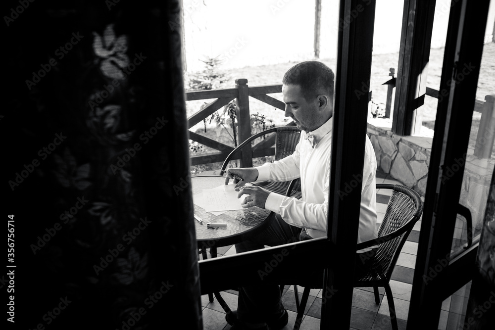 Obraz premium Black and white photo the groom in a shirt sits at a table on the terrace and writes a letter to the bride