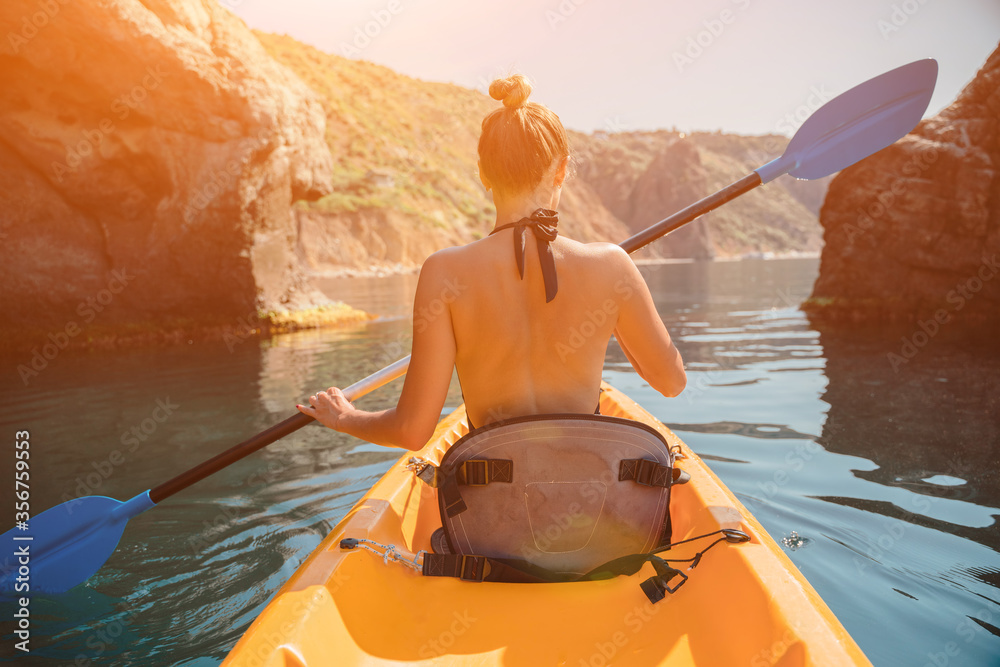 A girl in a swimsuit swims on a yellow kayak with a blue paddle in the sea. Against the backdrop of rocks and rocky shore. The concept of recreation, tourism, travel. Bright sunny day.