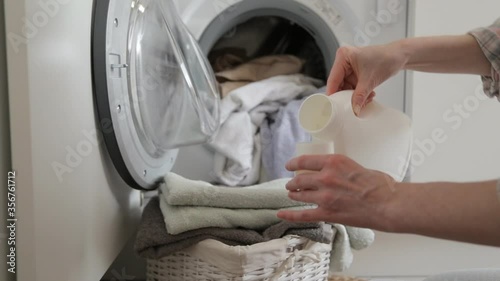 Woman pouring detergent gel into cap and put into washer machine, close the door and start washer. Washer machine and clothes with wicker basket in background