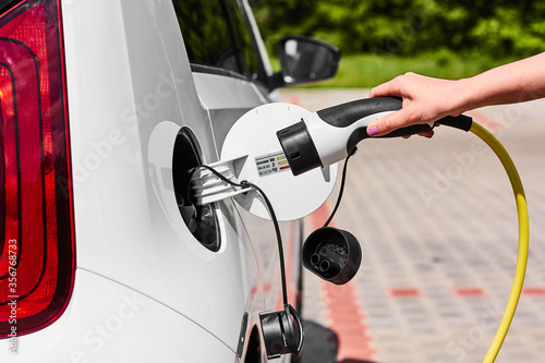 Close up woman hands plugging a power supply cable to electric car for charging at charging station outdoor. © vladim_ka